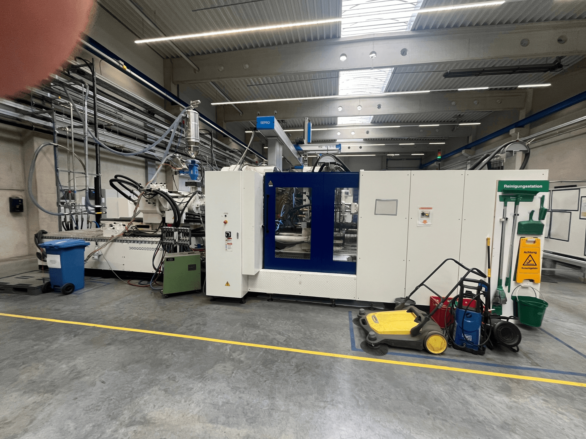 Průmyslový stroj v továrně, pohled zepředu, s modrými a bílými panely, hadicemi a čisticím zařízením poblíž.