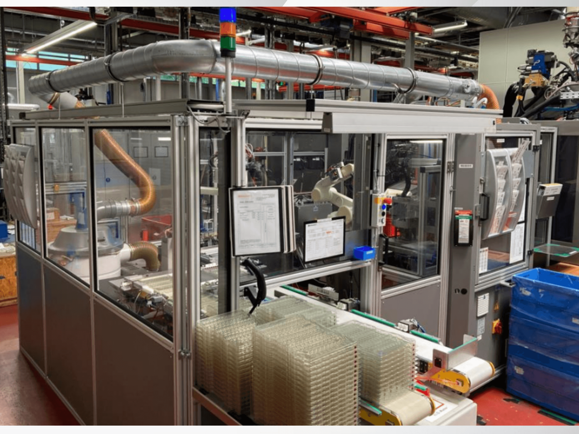 Pohled zepředu na automatizovaný stroj s kovovými trubkami, ovládacím panelem a naskládanými průhlednými plastovými nádobami na podlaze továrny.