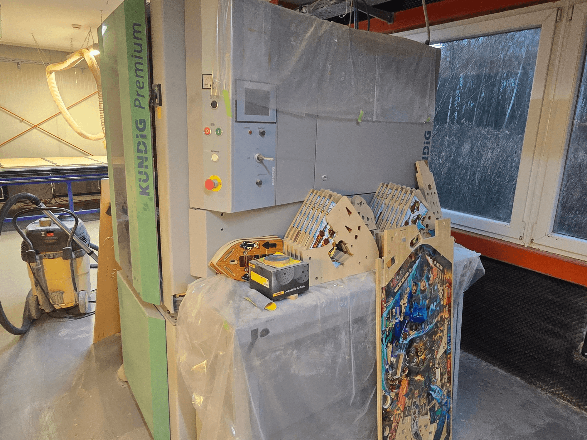 Pásová bruska Kundig Premium-1 1100-C v dílně, ovládací panel a kusy dřeva, boční pohled, s vysavačem prachu v pozadí.