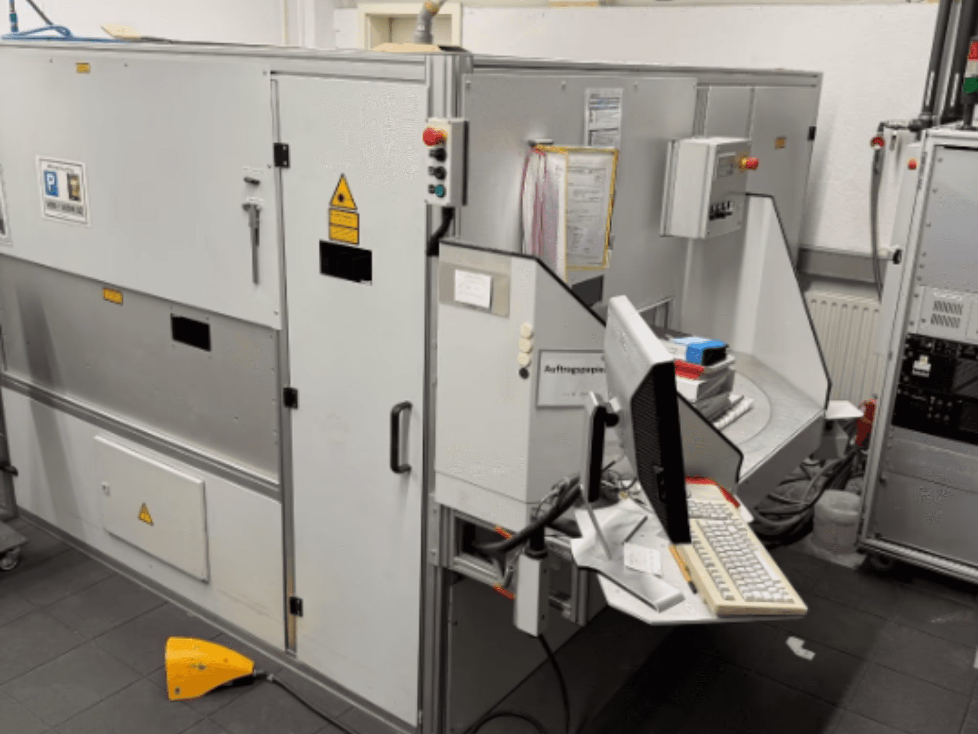 Průmyslový stroj s počítačovým terminálem, pohled z boku; včetně ovládacích tlačítek a bezpečnostních výstražných značek.