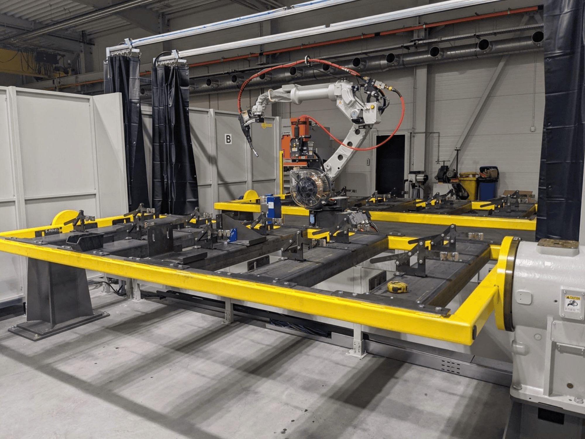 Pohled zepředu na robotické rameno PANASONIC TAWERS TM-2000WG3 umístěné nad žlutým pracovním stolem s kovovými díly určenými k montáži.