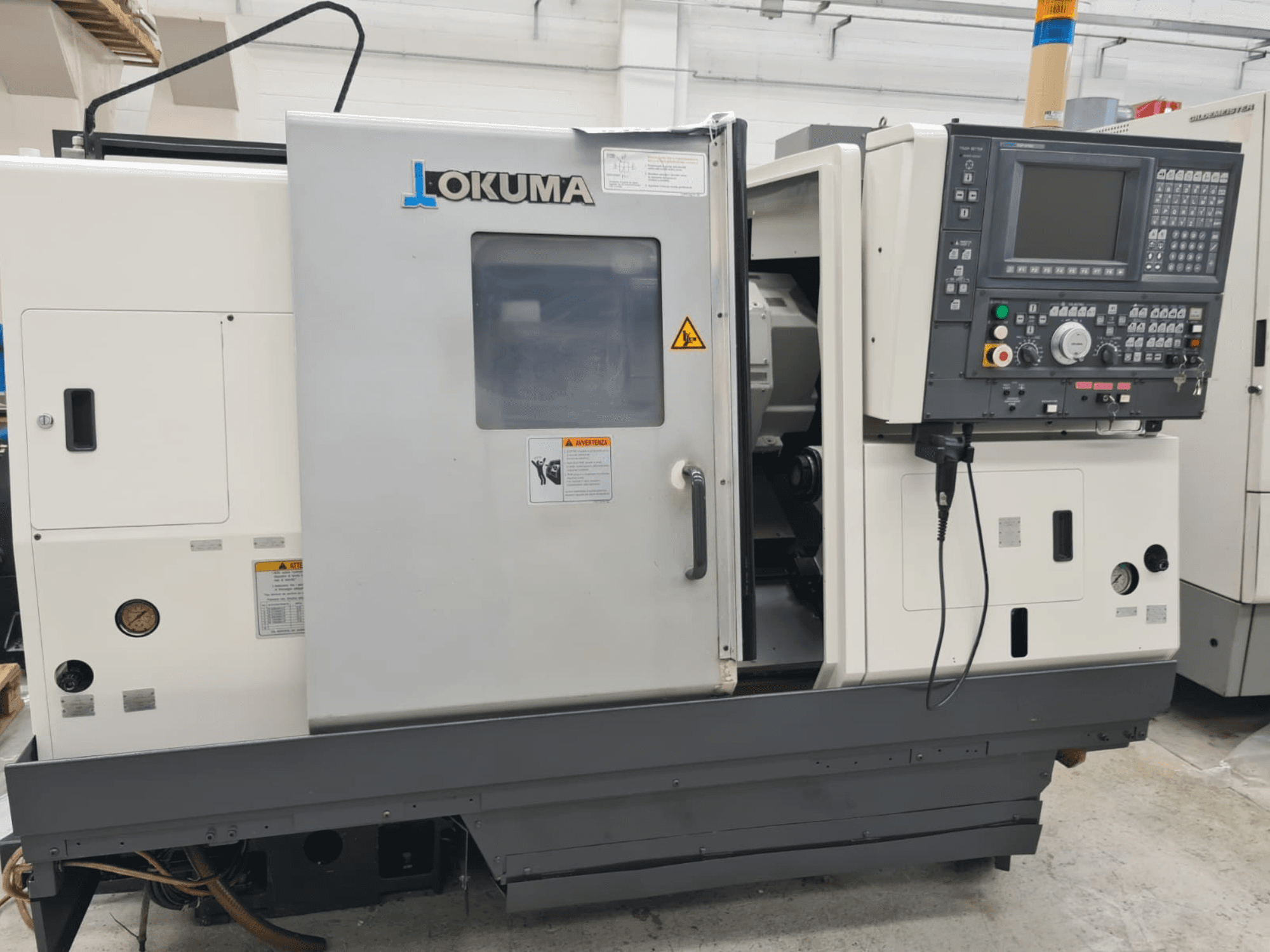Horizontální soustruh Okuma LB 300M X 500 v bočním pohledu, na kterém je vidět ovládací panel, přístup k vřetenu a bezpečnostní upozornění.