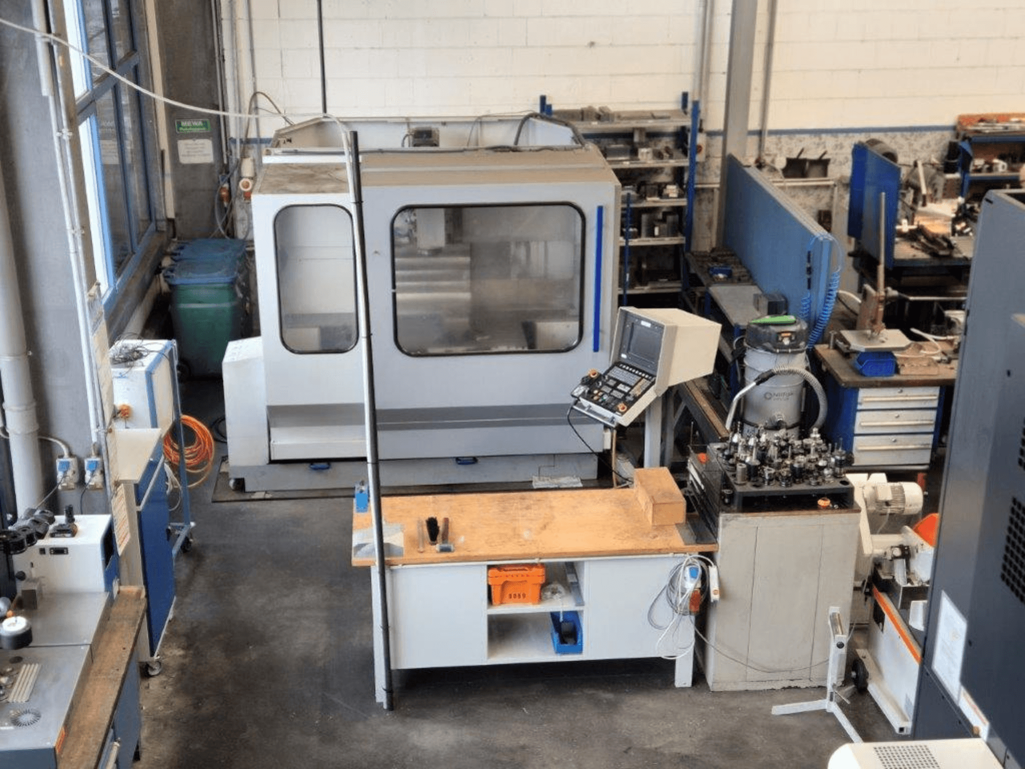 CNC frézka v dílně, pohled zepředu, v pozadí je vidět ovládací panel, pracovní plocha a nástroje.