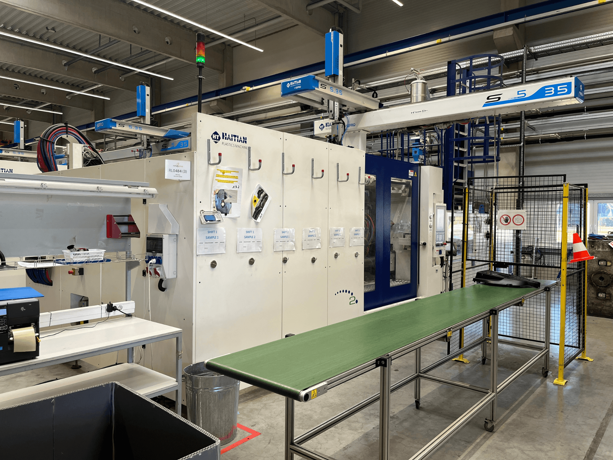 Haitský stroj na zpracování plastů S5-35 v dílně; zepředu je vidět zelený dopravní stůl, bezpečnostní zábrany a ovládací panel.