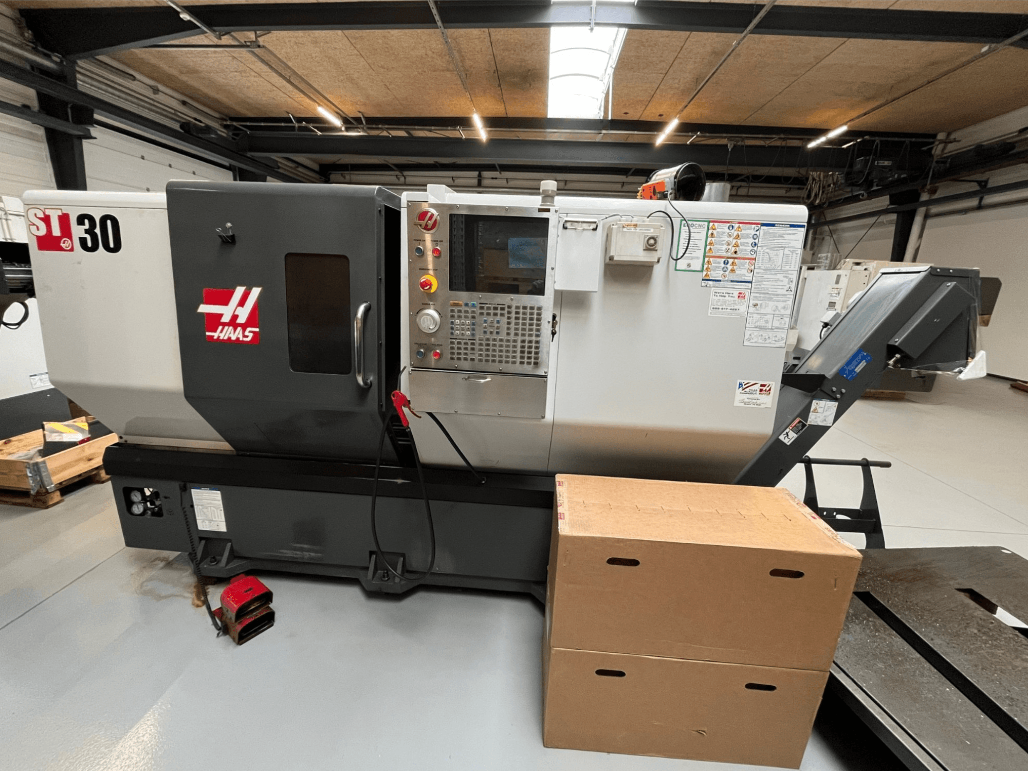CNC soustruh Haas ST-30, pohled zepředu, ovládací panel s tlačítky, nouzové zastavení a dvě na sebe naskládané kartonové krabice.