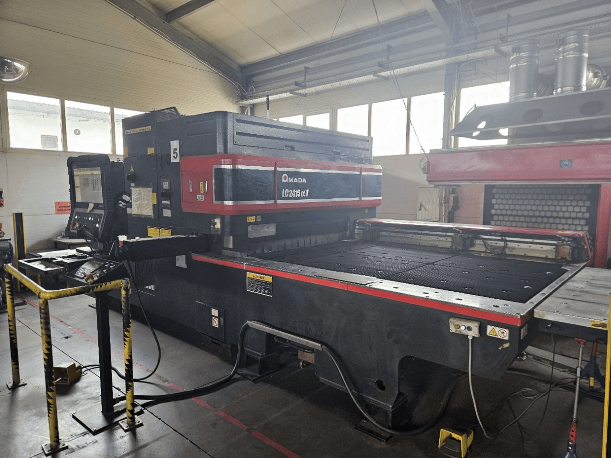 Laserový řezací stroj AMADA LC2415 Alpha 5 s CO₂ laserem, pohled zepředu znázorňující ovládací panel a řezací stůl s bezpečnostními zábranami.