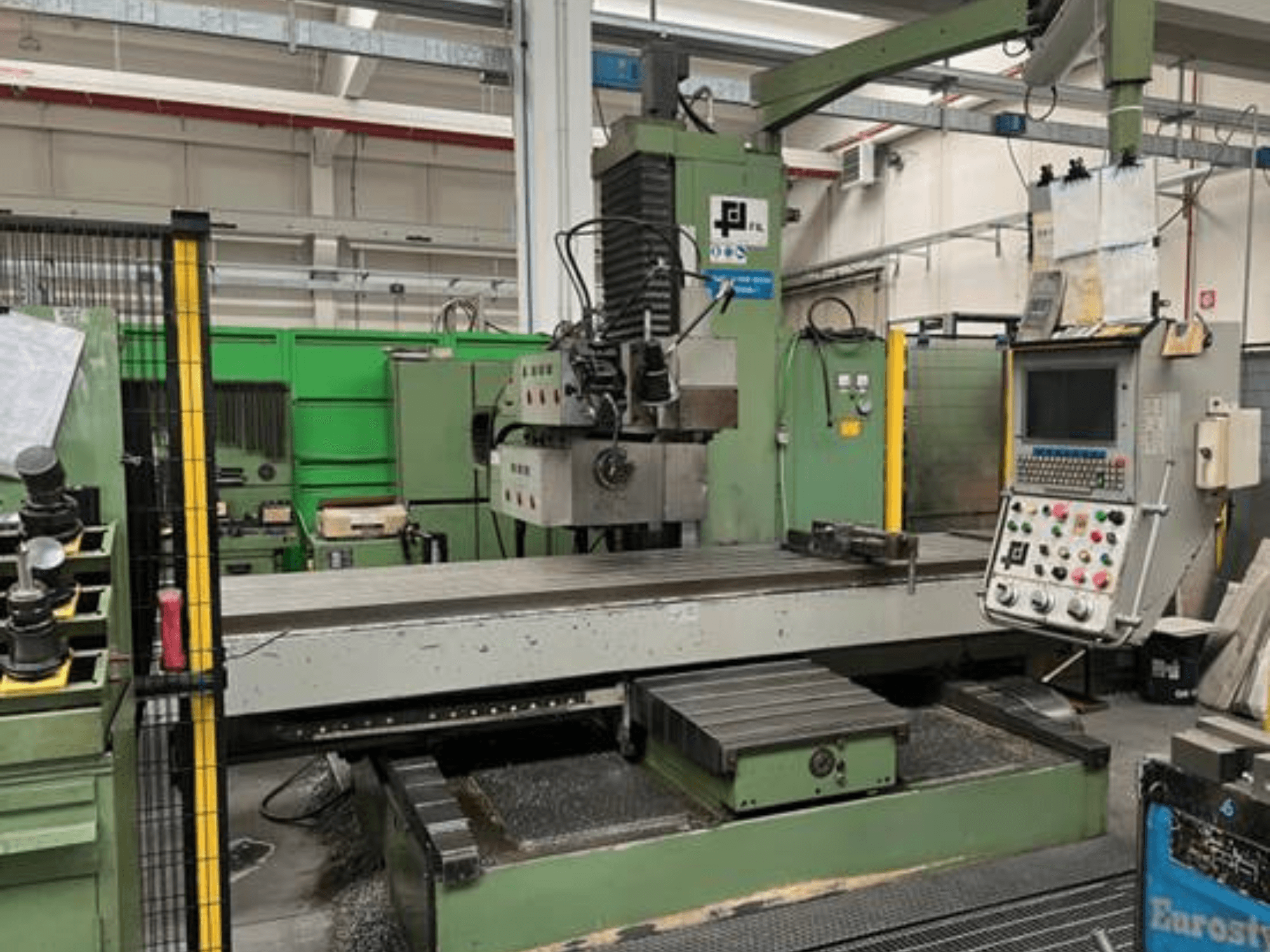 Průmyslová frézka od společnosti JFL, pohled zepředu s ovládacím panelem, velkým plochým stolem a kovovými hoblinami na podlaze.