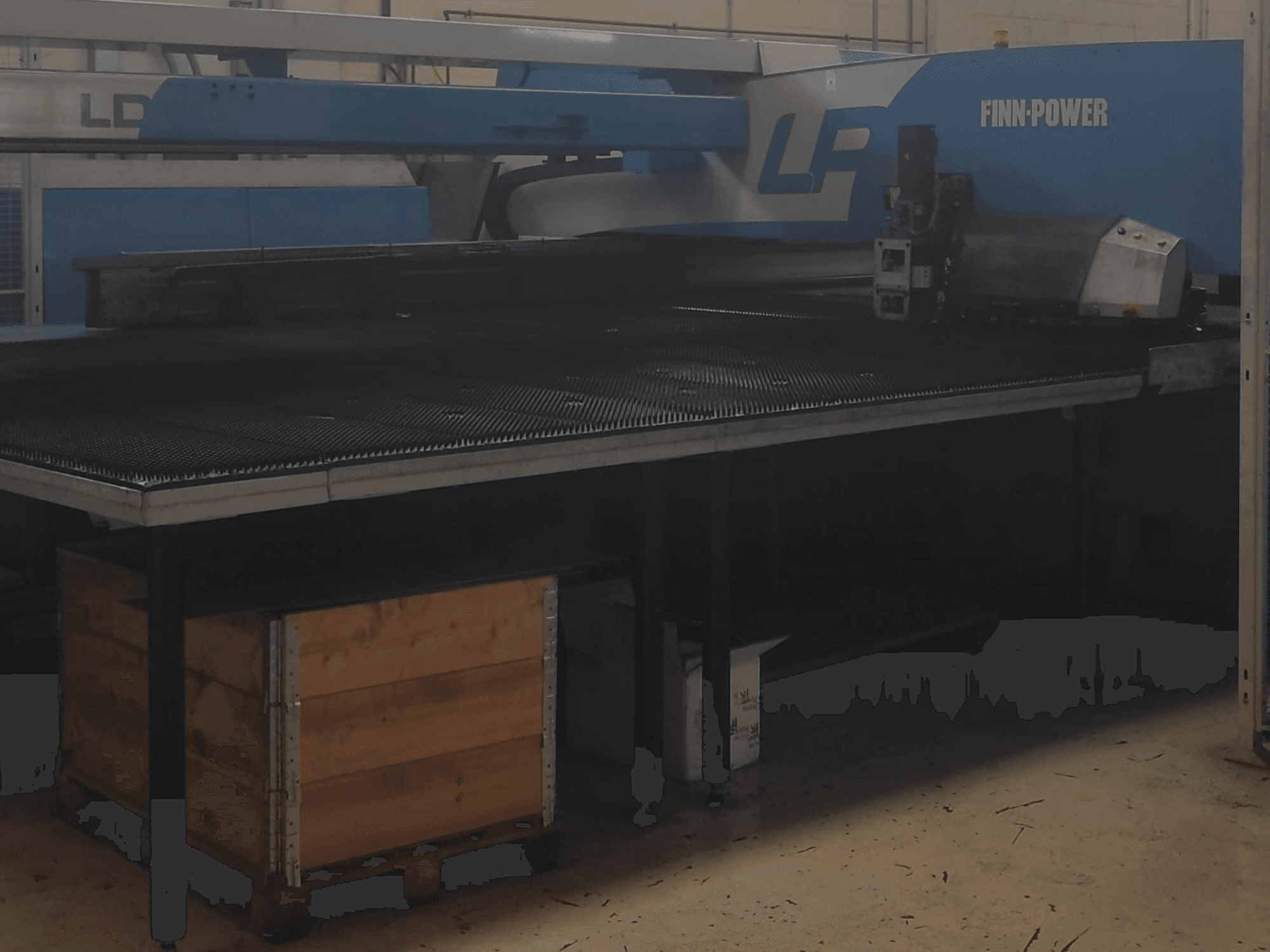 Řezací stroj Finn-Power LP v továrně, pohled zepředu s kovovým stolem s texturou a dřevěnou bednou pod ním.