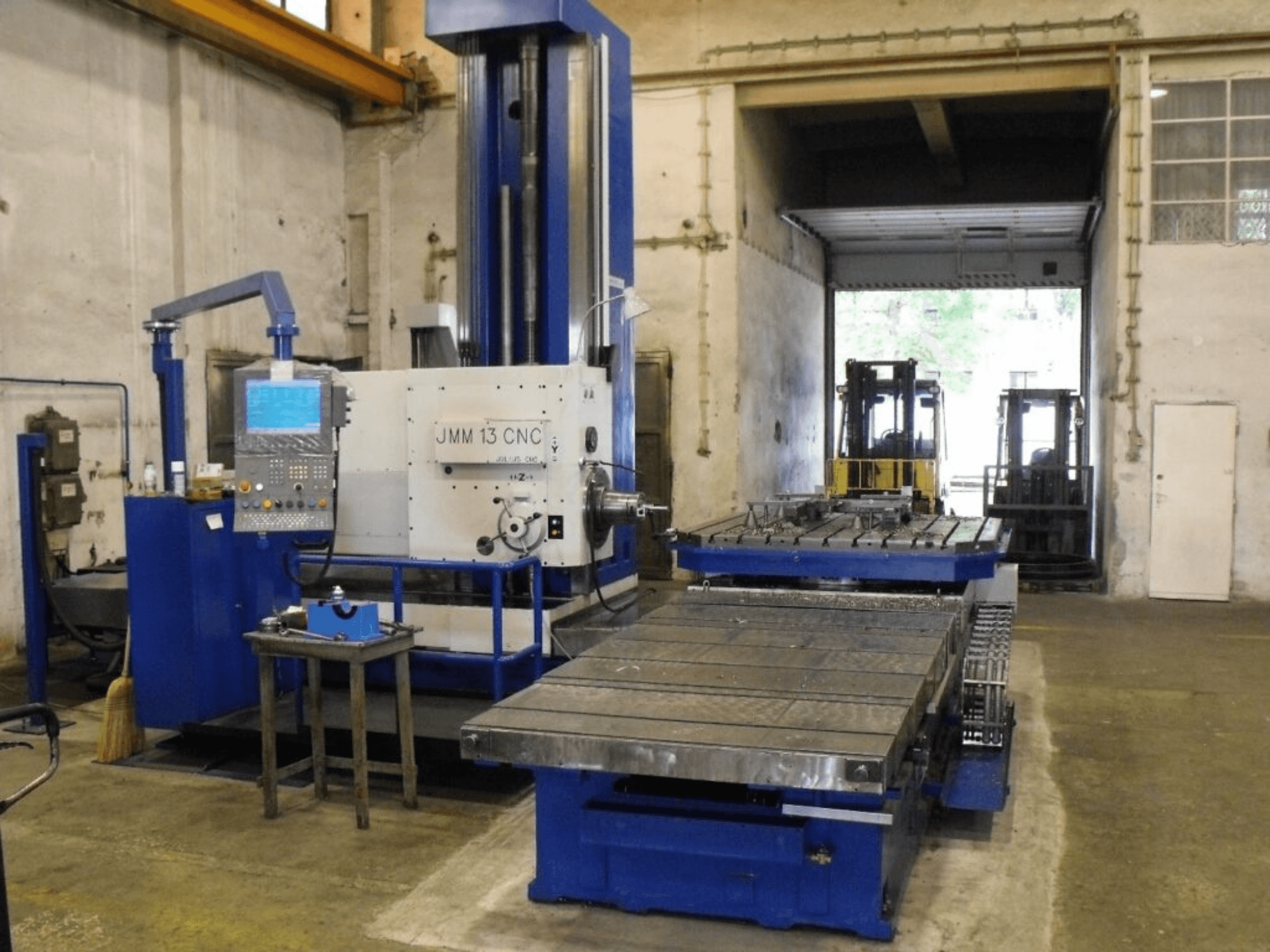 Frézka JMM 13 CNC v dílně, pohled zepředu, s ovládacím panelem, velkým pracovním stolem a průmyslovým pozadím.