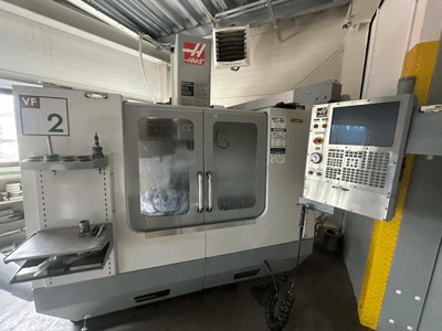 CNC stroj Haas VF-2, pohled zepředu, s ovládacími prvky, držákem nástrojů a průhlednými dvířky s viditelnými nástroji uvnitř skříně.