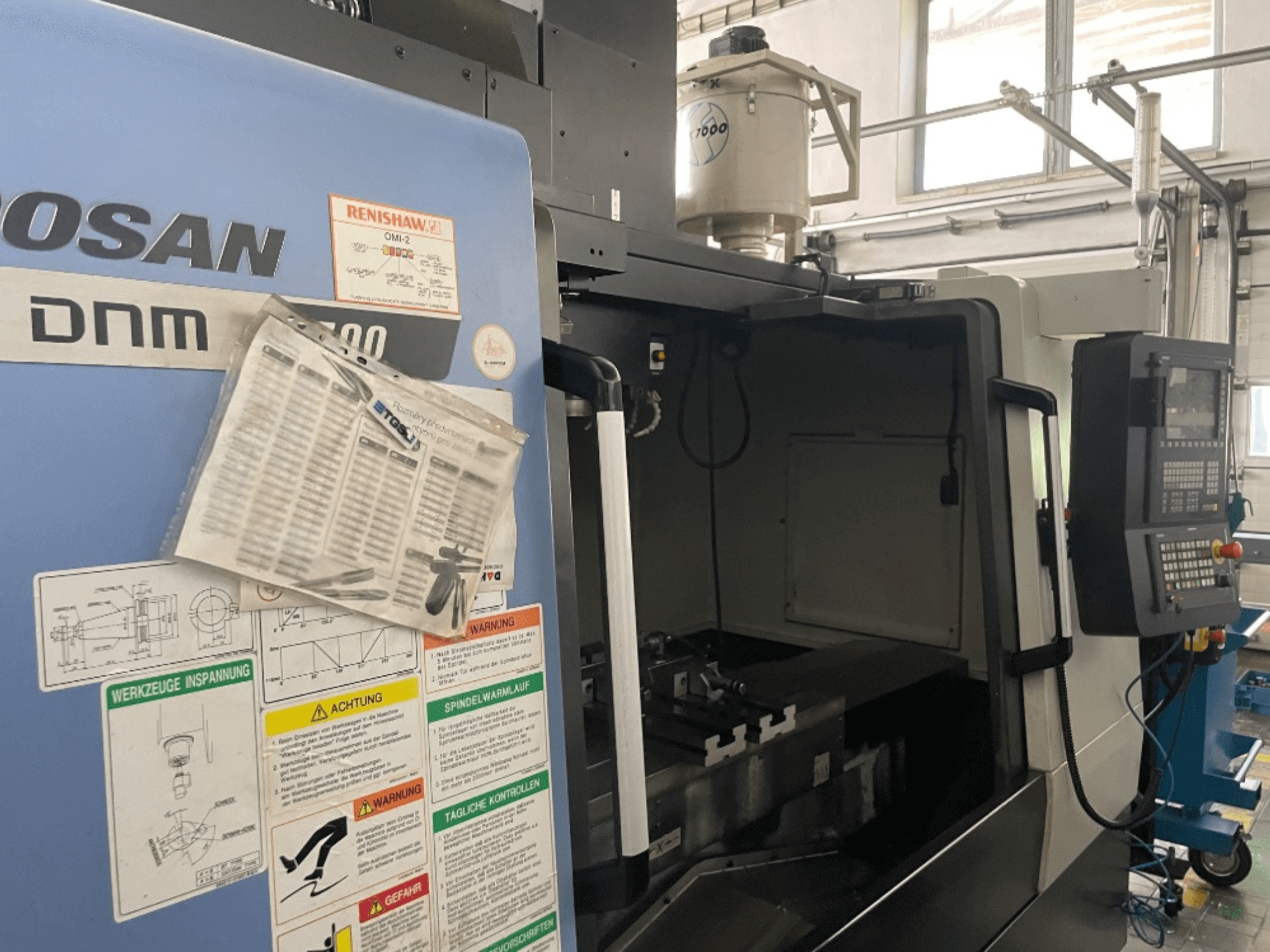Vertikální obráběcí centrum DOOSAN DNM 5700, pohled zepředu, zobrazuje ovládací panel, bezpečnostní štítky a detaily stroje.