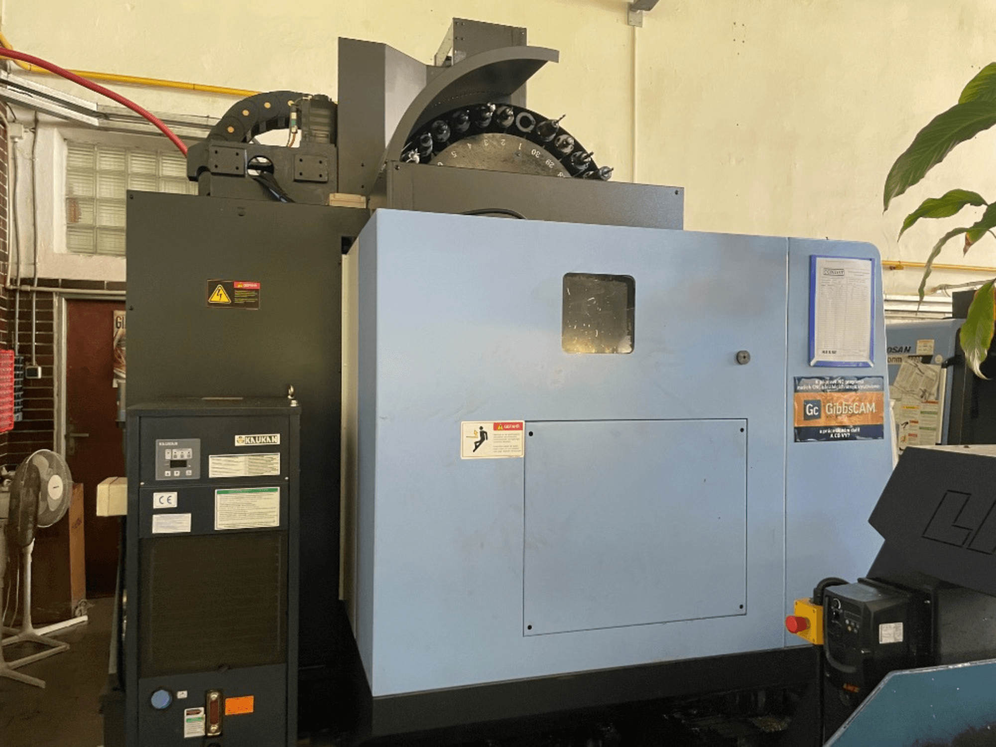 Pohled zepředu na vertikální obráběcí centrum DOOSAN DNM 5700, zobrazující ovládací panel, bezpečnostní štítky a funkce stroje.