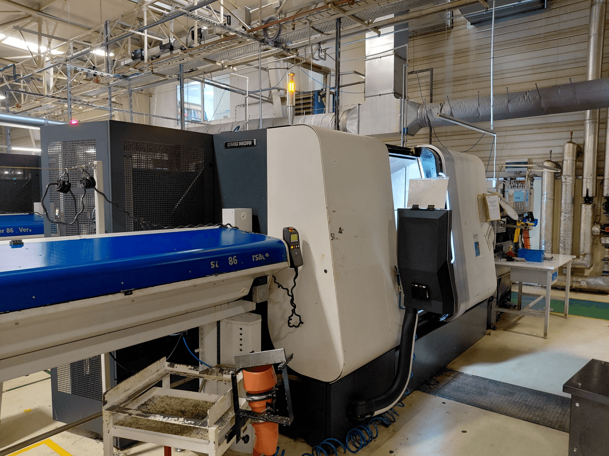 Lineární vícevřetenový soustruh DMG MORI Sprint 65, boční pohled s viditelným ovládacím panelem a dopravníkem v dílenském prostředí.