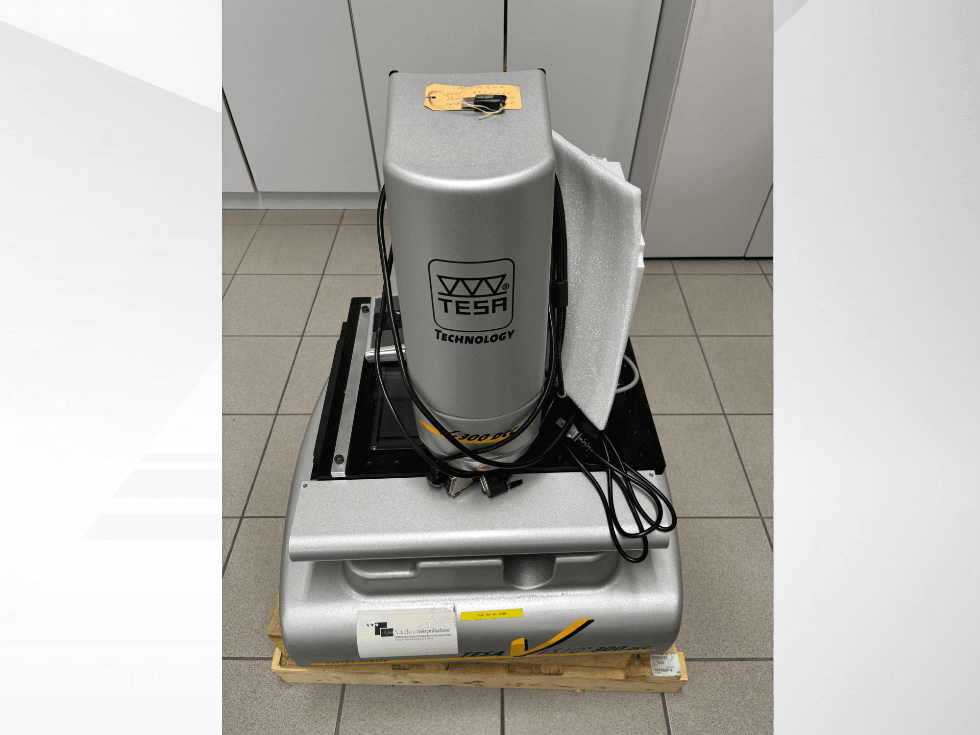 Optický měřicí stroj TESA Visio 300 DCC v čelním pohledu s kabely, na dřevěné paletě a s viditelným ochranným obalem.