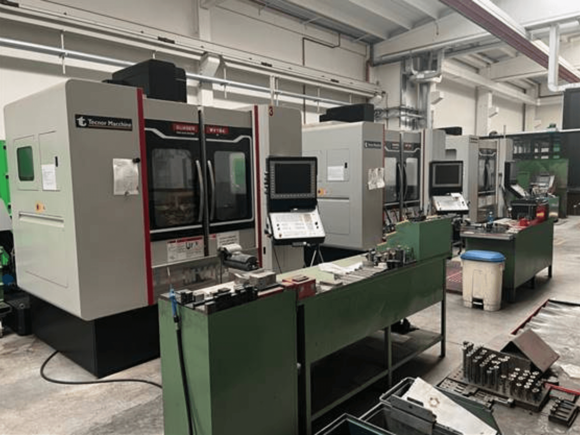 Čtyři CNC stroje Tecnoer Marchine v dílně, pohled zepředu, s nástroji a díly na zelených pracovních stolech.
