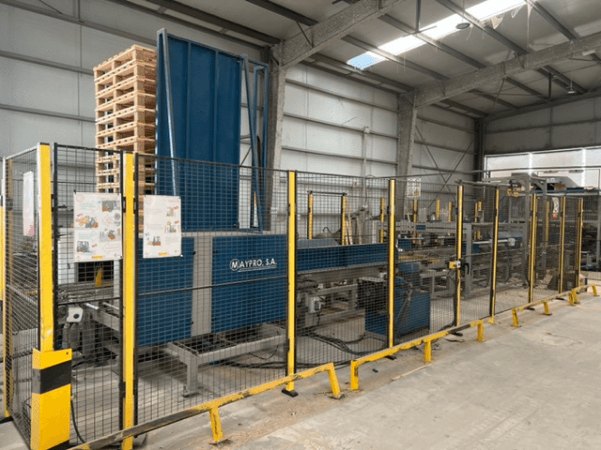 Průmyslový balicí stroj na palety od společnosti Maypro S.A. ve skladu, s bezpečnostní sítí a ovládacími prvky, boční pohled.