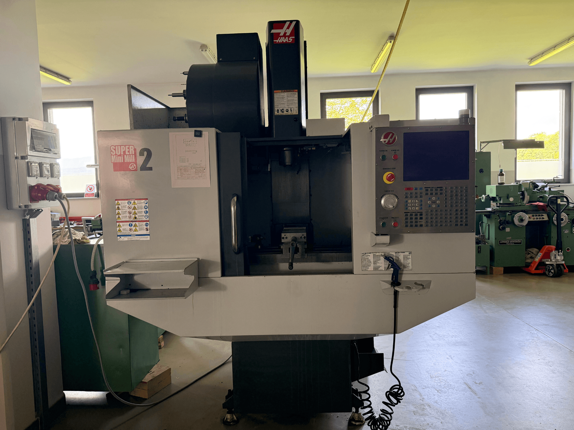 Vertikální obráběcí centrum Haas Mini Mill 2 v čelním pohledu, na kterém je vidět ovládací panel a prostor pro nástroje s výstražnými štítky.