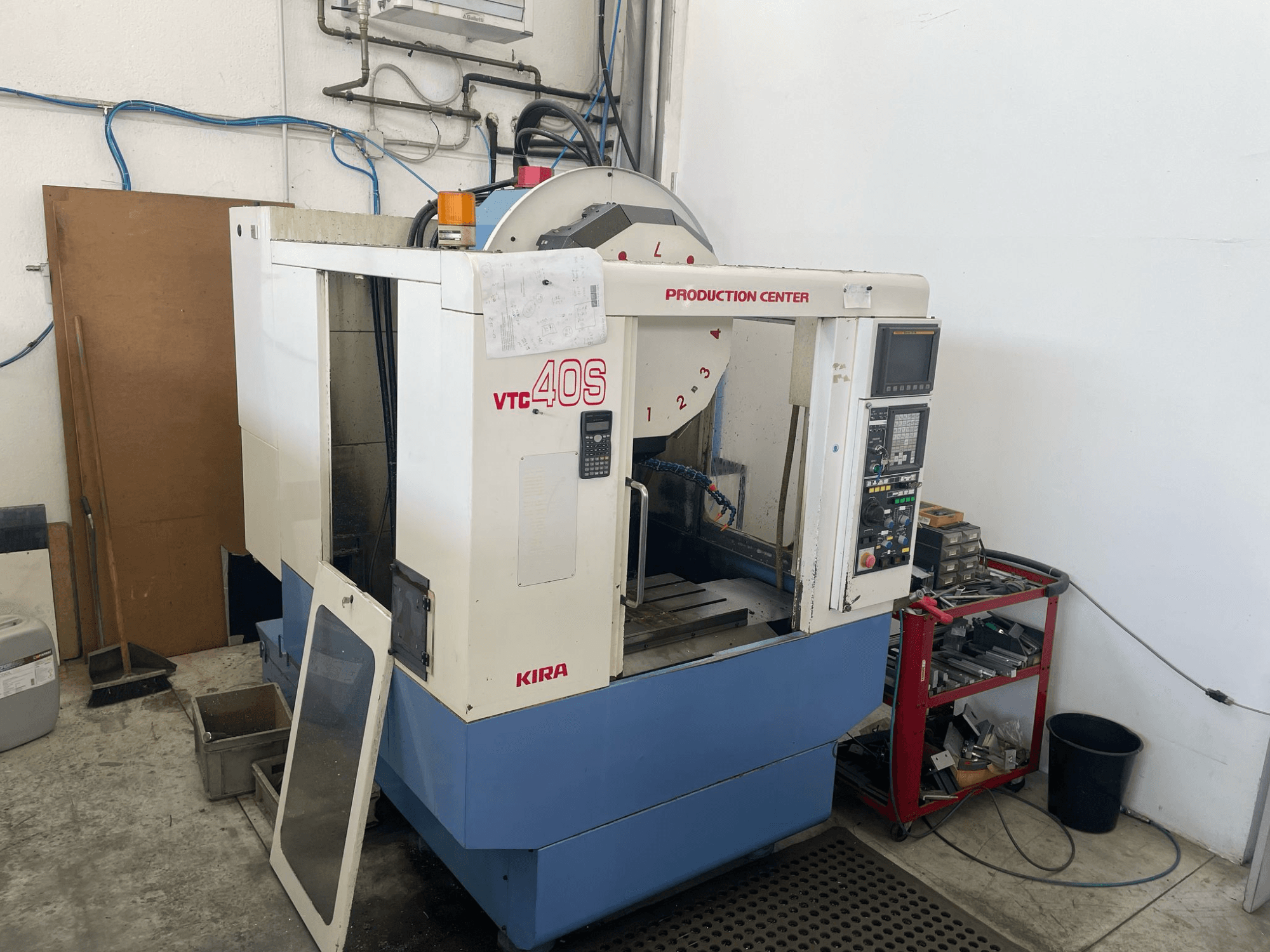 Obráběcí centrum KIRA VTC 40S v bočním pohledu, s ovládacím panelem, otevřenými dvířky a nástroji na úložném vozíku poblíž.