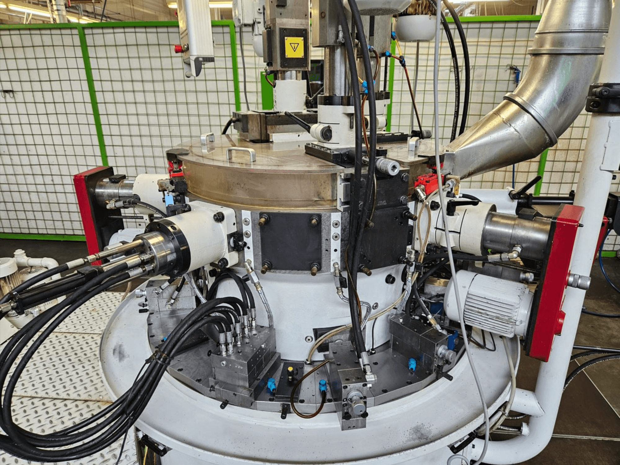 Detailní pohled na průmyslový stroj s hydraulickými hadicemi, kovovými součástmi a rotující základnou. Značka není viditelná.