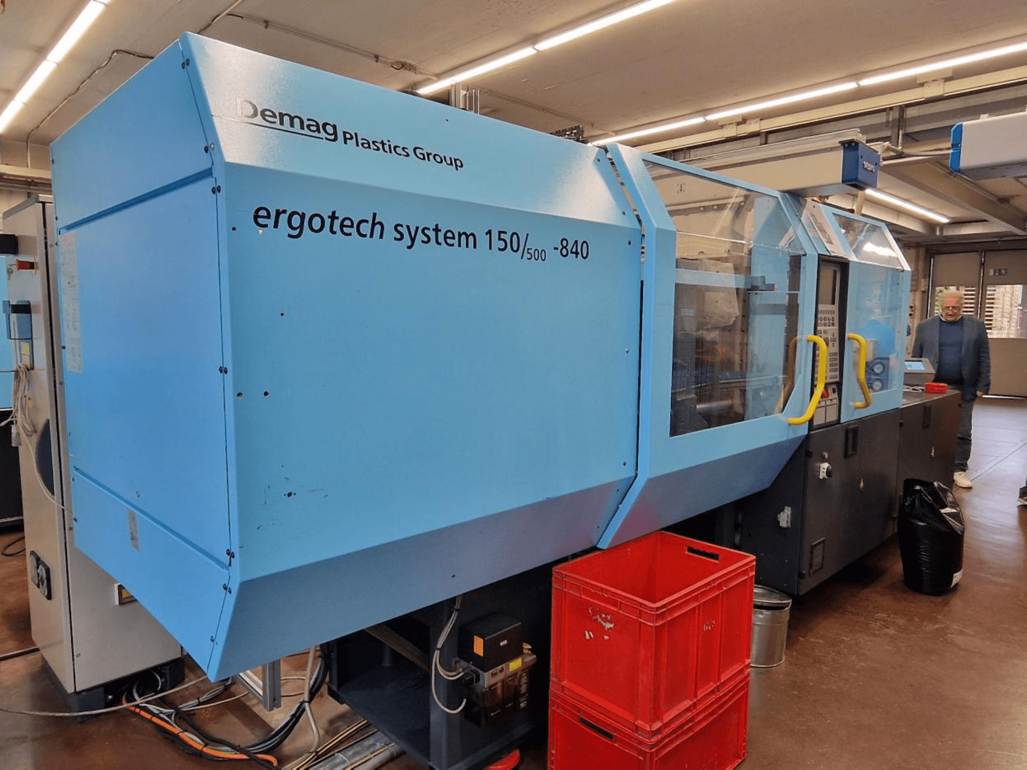 Vstřikovací stroj Demag Ergotech System 150/500-840 v modré barvě, boční pohled, s bednami a obsluhou v pozadí.