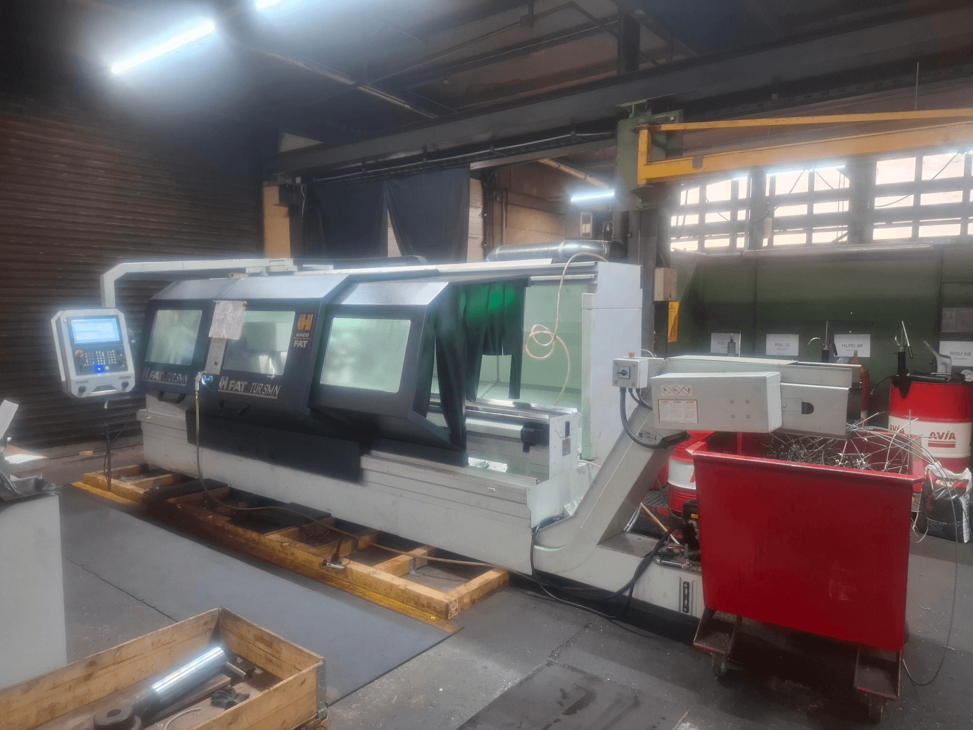 Pohled zepředu na CNC stroj FAT Tursan s ovládacím panelem, umístěný na dřevěných paletách v dílně.