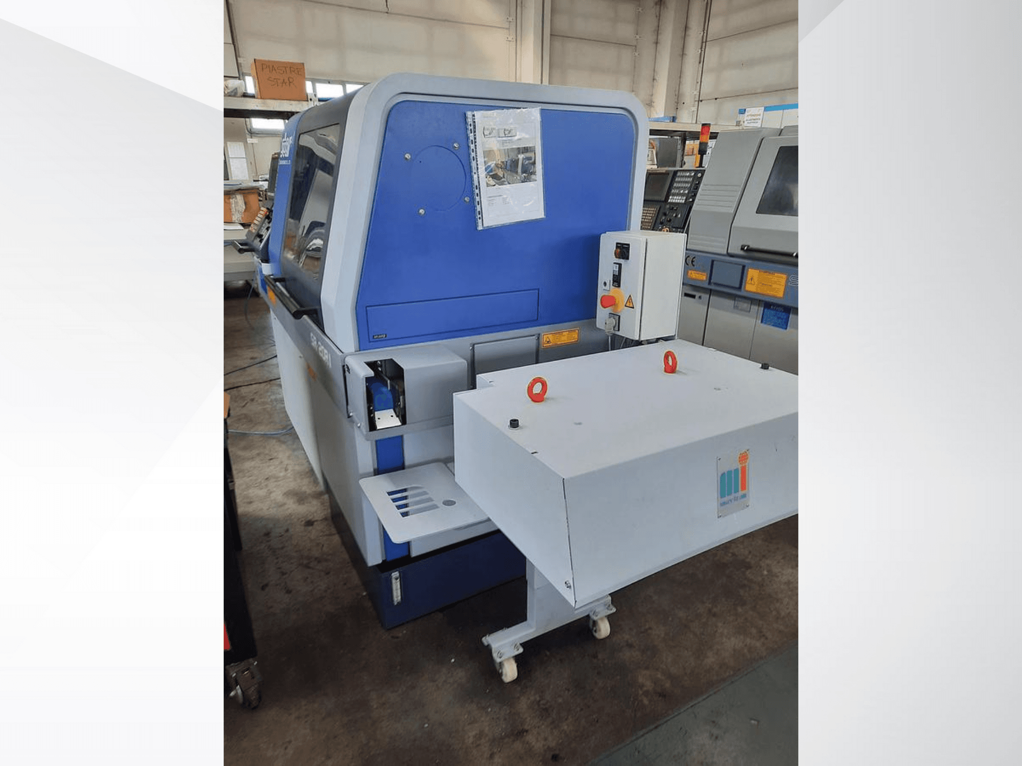 Modrý průmyslový stroj s bílým dopravníkem, zobrazující ovládací panel a připojené bezpečnostní prvky, boční pohled.
