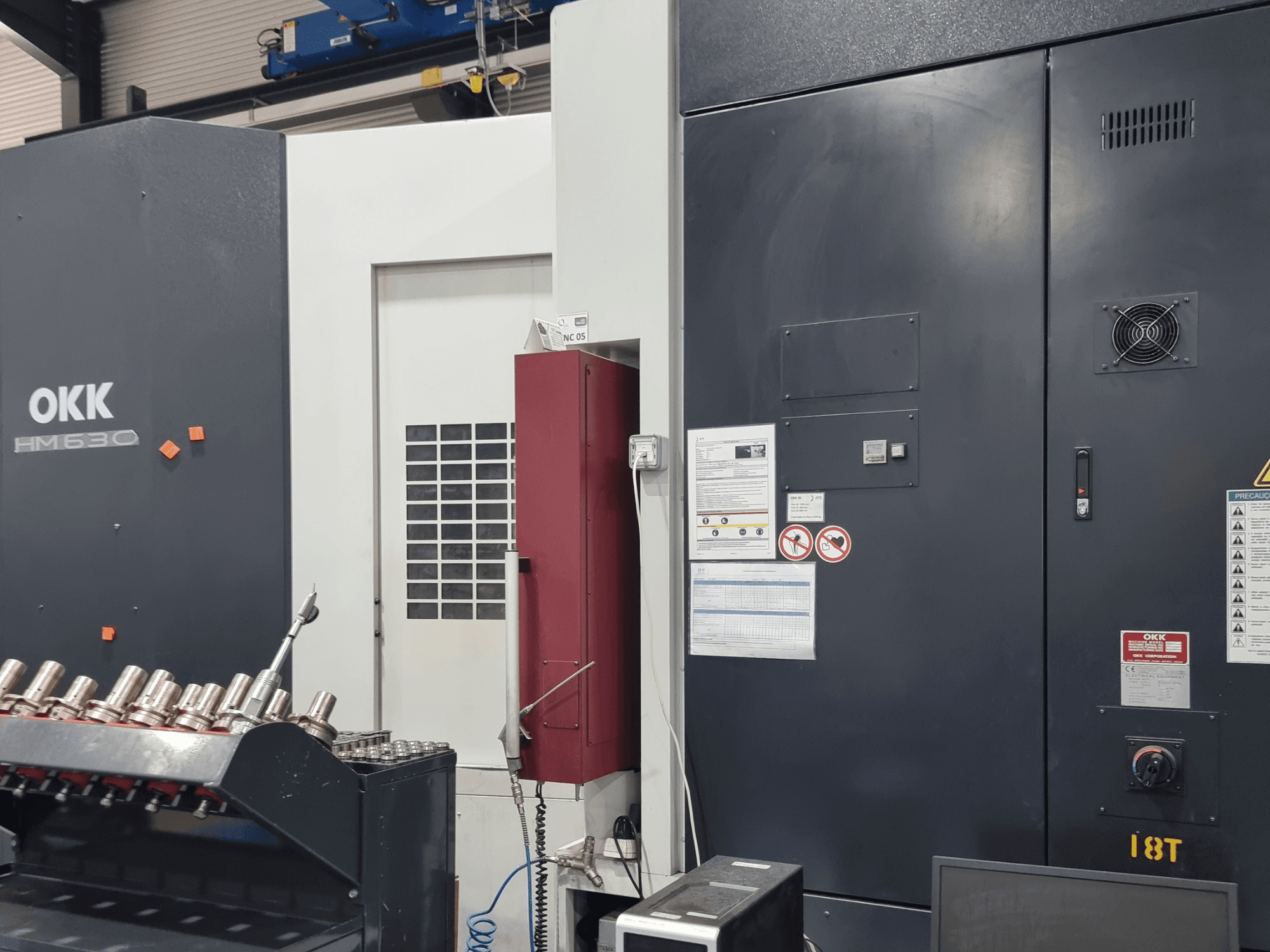 Horizontální obráběcí centrum OKK HM630, pohled zepředu znázorňující ovládací panel, bezpečnostní pokyny a úložný prostor pro nástroje na boku.