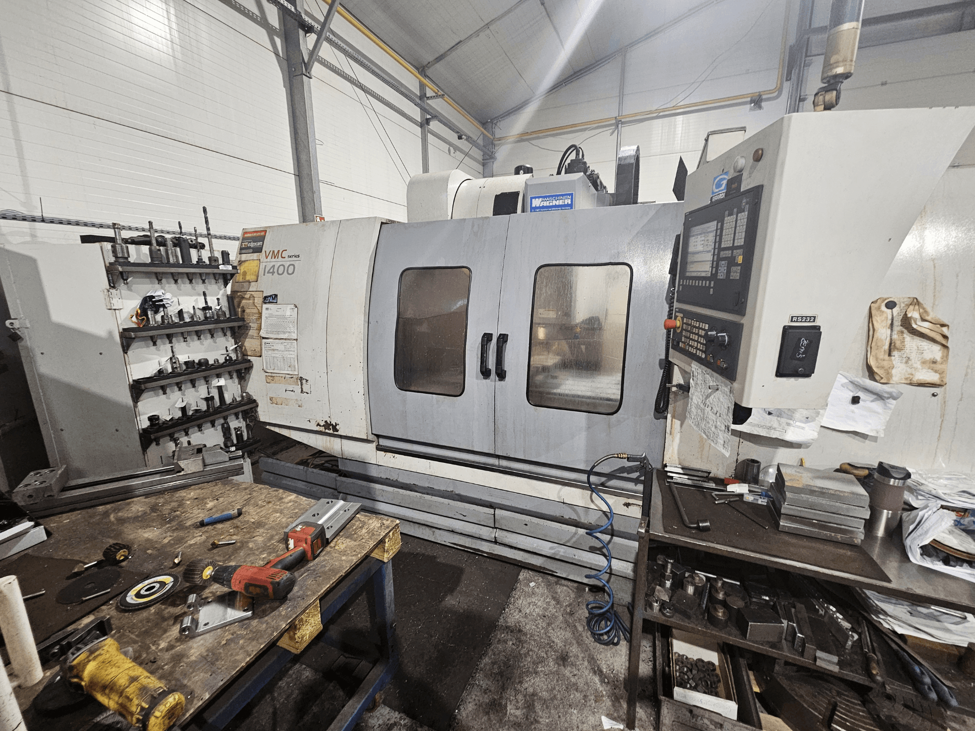 Frézka VMC 1400 CNC v dílně, pohled zepředu; nástroje a materiály rozptýlené na pracovním stole poblíž.