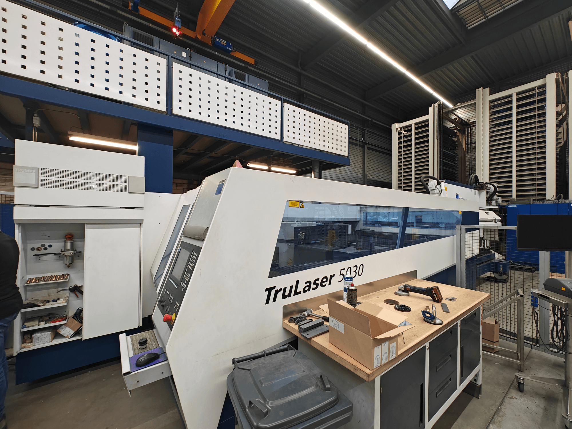 Stroj TruLaser 5030 v dílně, pohled zepředu s viditelným ovládacím panelem, obklopený nástroji a zařízením.