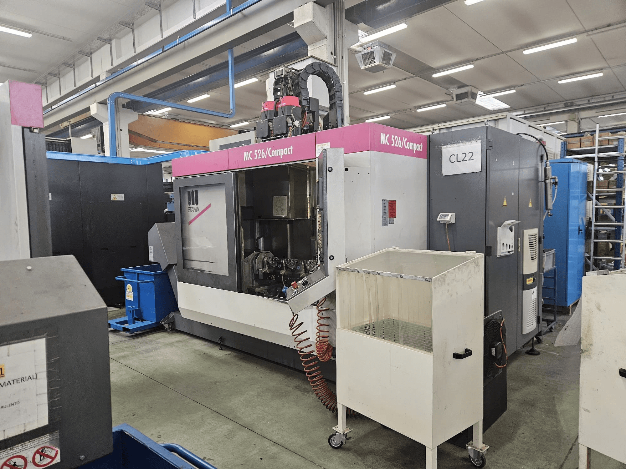 Kompaktní CNC stroj MC 526 od společnosti Stama, boční pohled, zobrazující ovládací panel, nádrž chladicí kapaliny a přilehlé stroje.