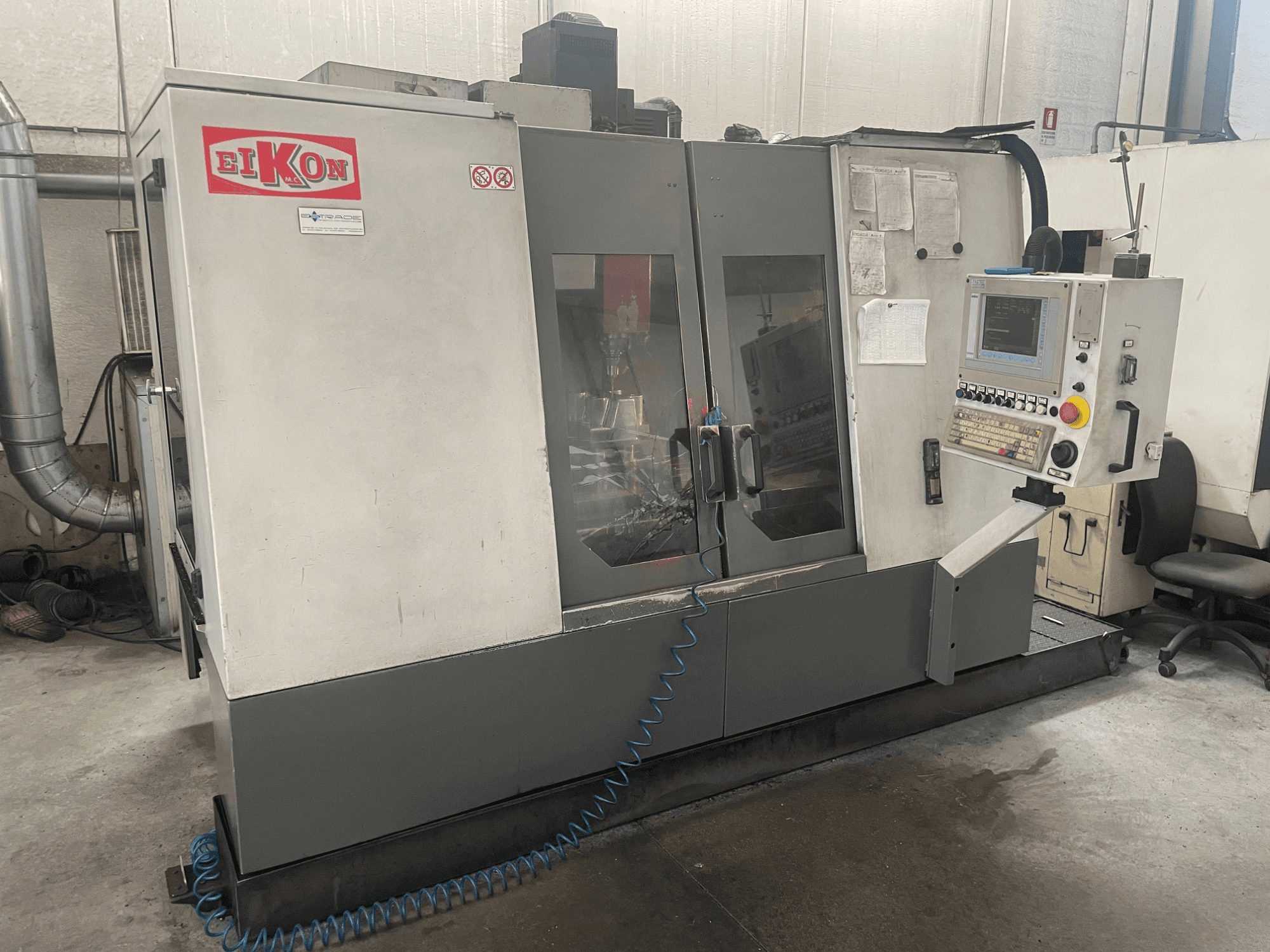 Stroj EIKON CNC při pohledu zepředu, s ovládacím panelem, velkými skleněnými dveřmi a modrou vzduchovou hadicí na podlaze.