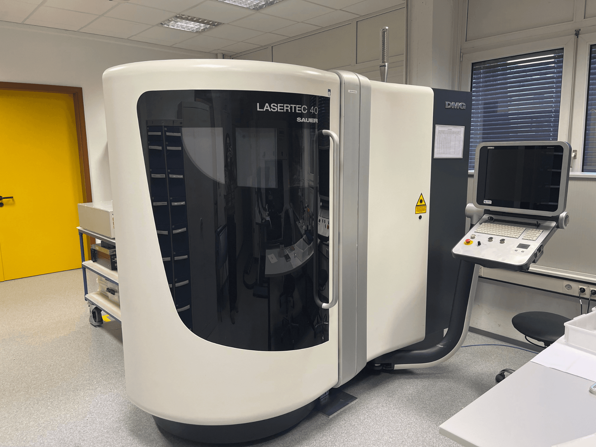 Bílá frézka LASERTEC 40 od DMG MORI, pohled zepředu, s ovládacím panelem a ergonomickým designem v dobře osvětlené místnosti.