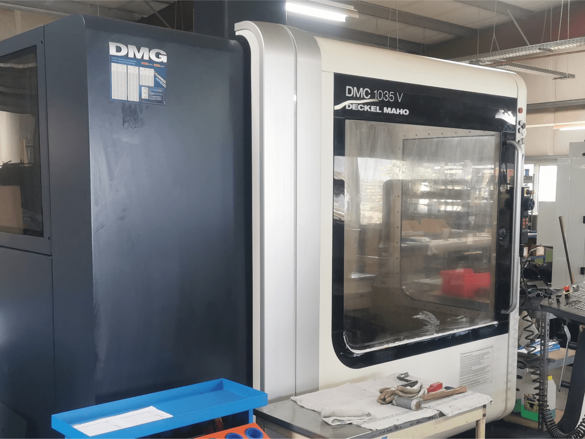 Frézka DMG DMC 1035 V v dílně, pohled zepředu s modrým zásobníkem nástrojů a rukavicemi na povrchu.