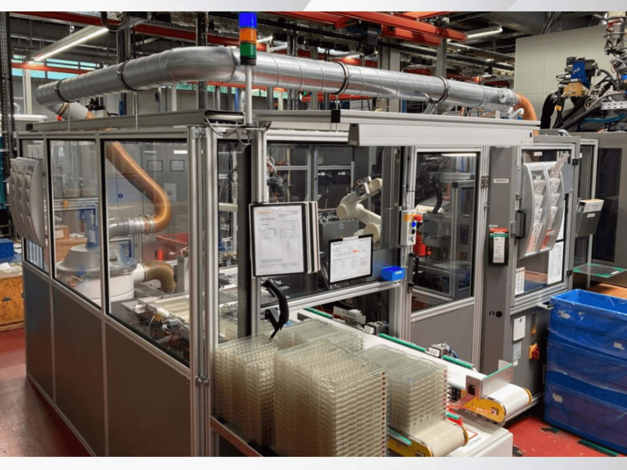 Automatizovaný montážní stroj s robotickým ramenem, průhlednými bočními panely a naskládanými kontejnery s průhlednými předměty, pohled zepředu.