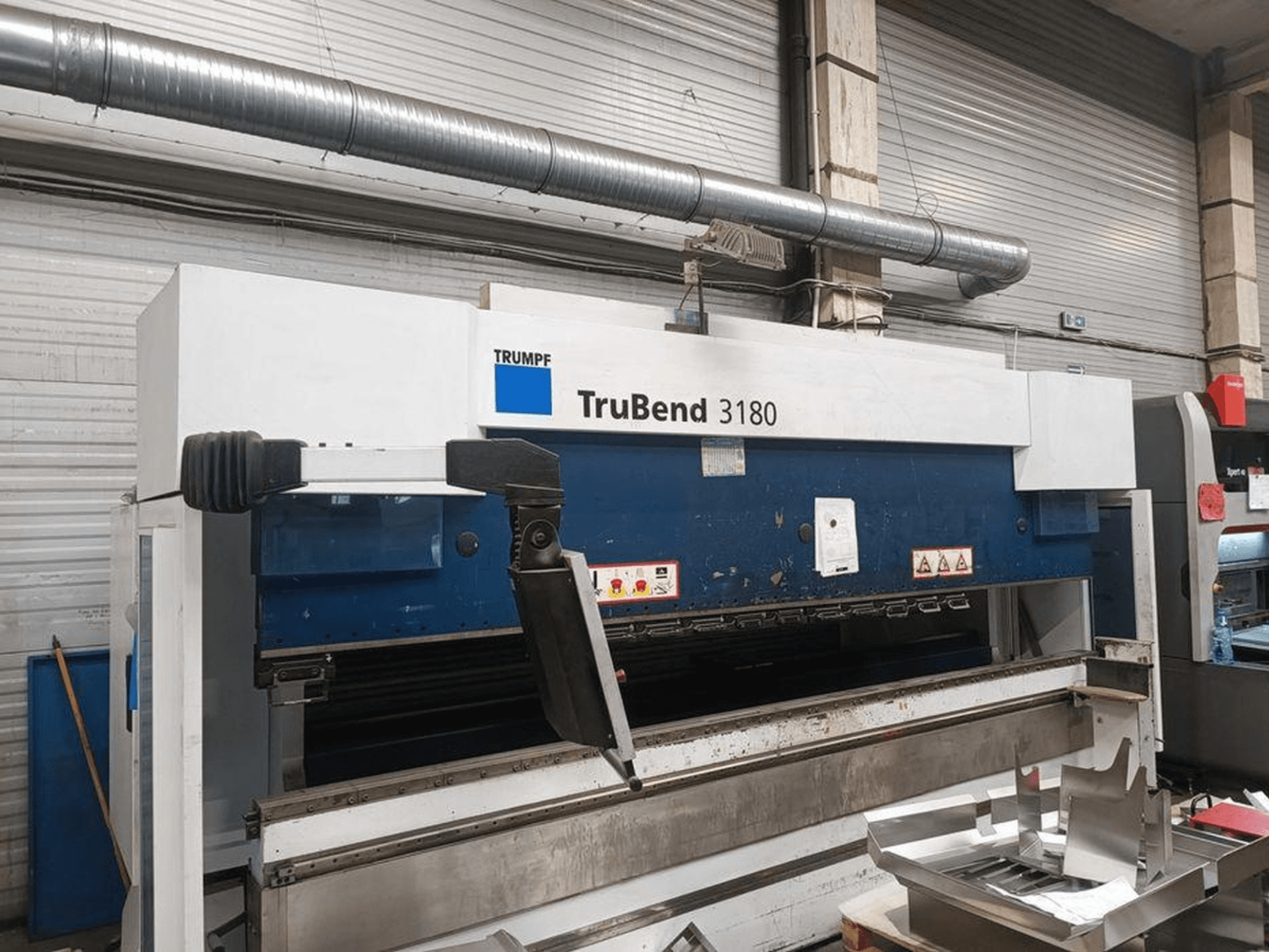 Ohýbačka Trumpf TruBend 3180 v dílně, boční pohled ukazuje ovládací panel a kovové kusy na stole.