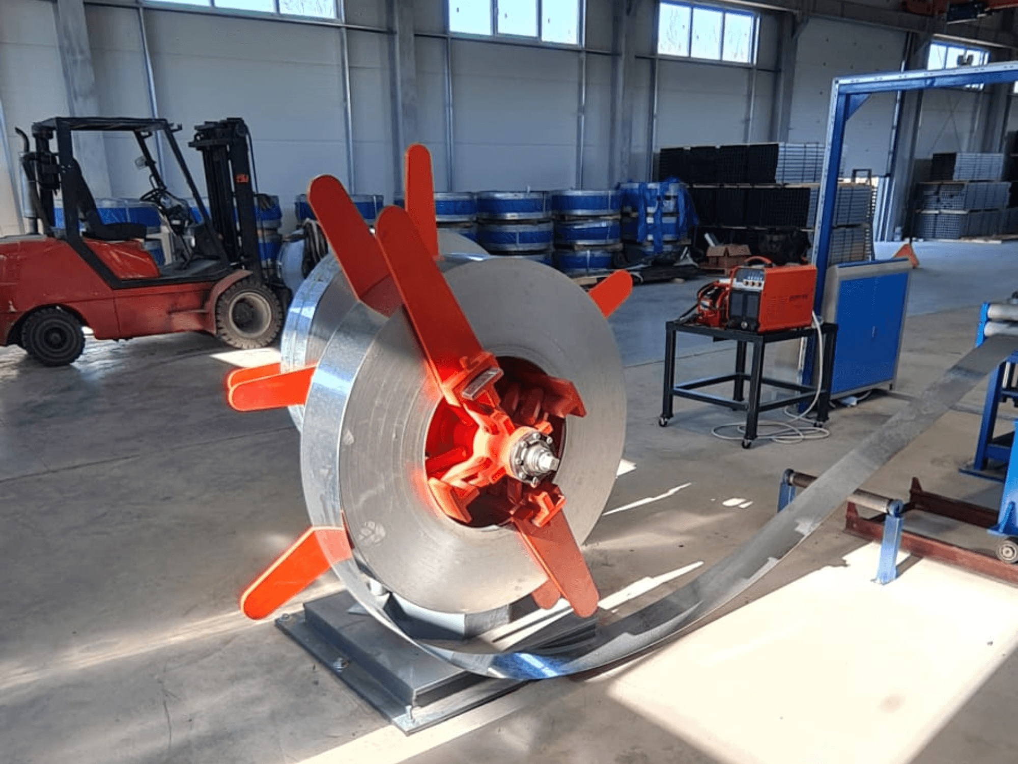 Kovová cívka s červenými úchyty na stojanu, pohled z úhlu. Průmyslové pozadí se stroji a vysokozdvižným vozíkem.