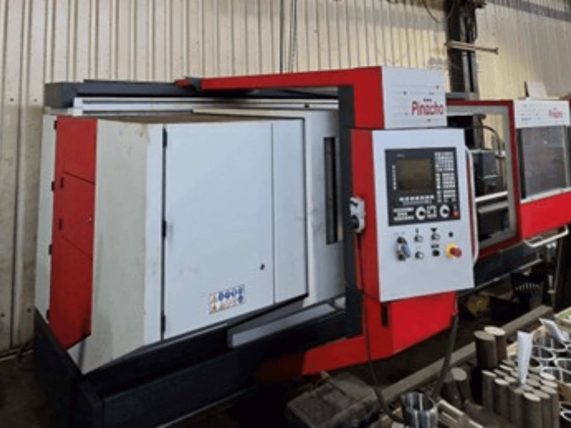 Stroj Pinacho CNC v červenobílé barvě, pohled z boku, s ovládacím panelem a materiály na podlaze, znázorňující provozní nastavení.