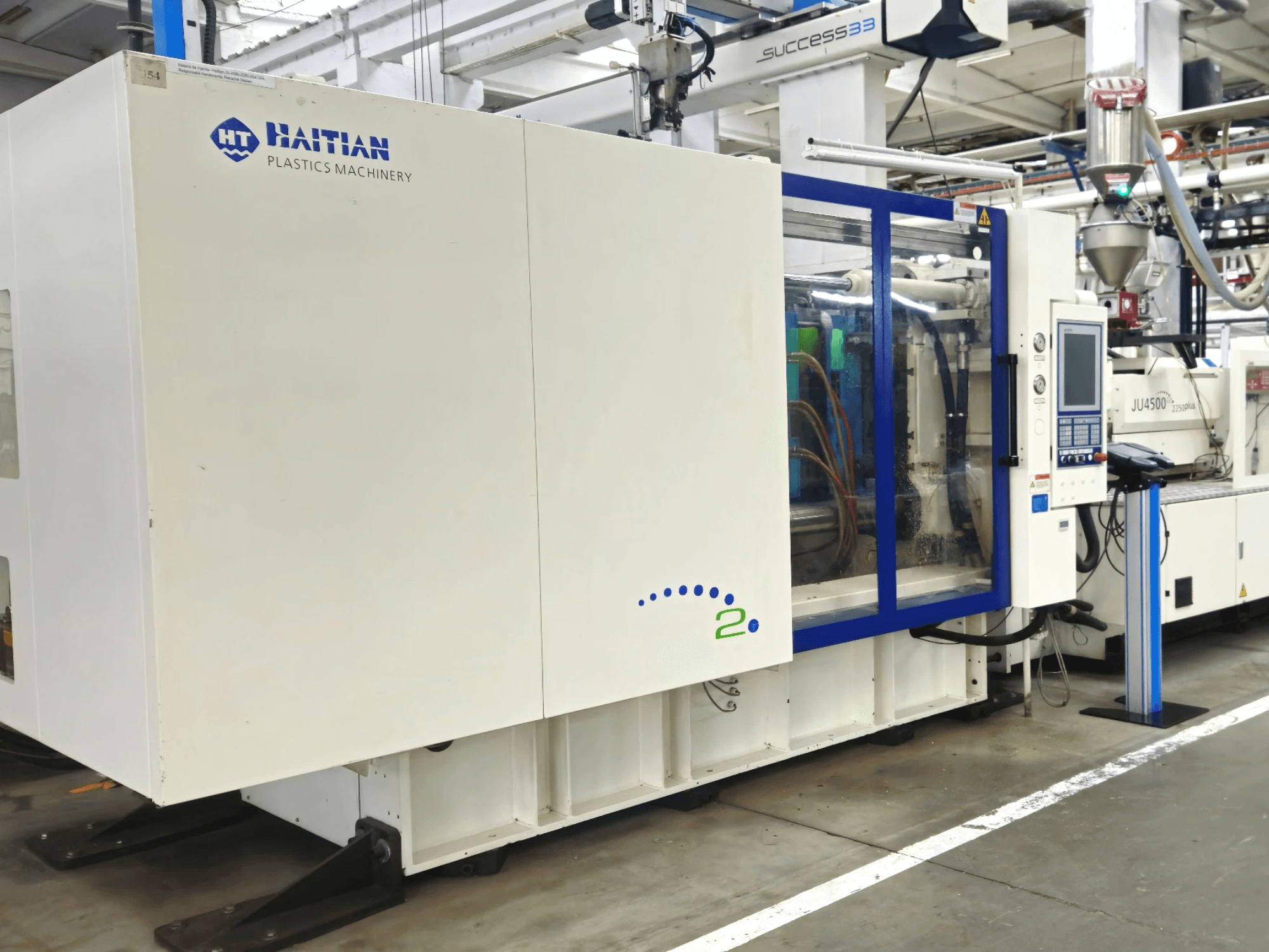 Model Haitian Plastics Machinery, pohled zepředu, představující strojní zařízení s ovládacím panelem a viditelnými trubkami v průmyslovém prostředí.