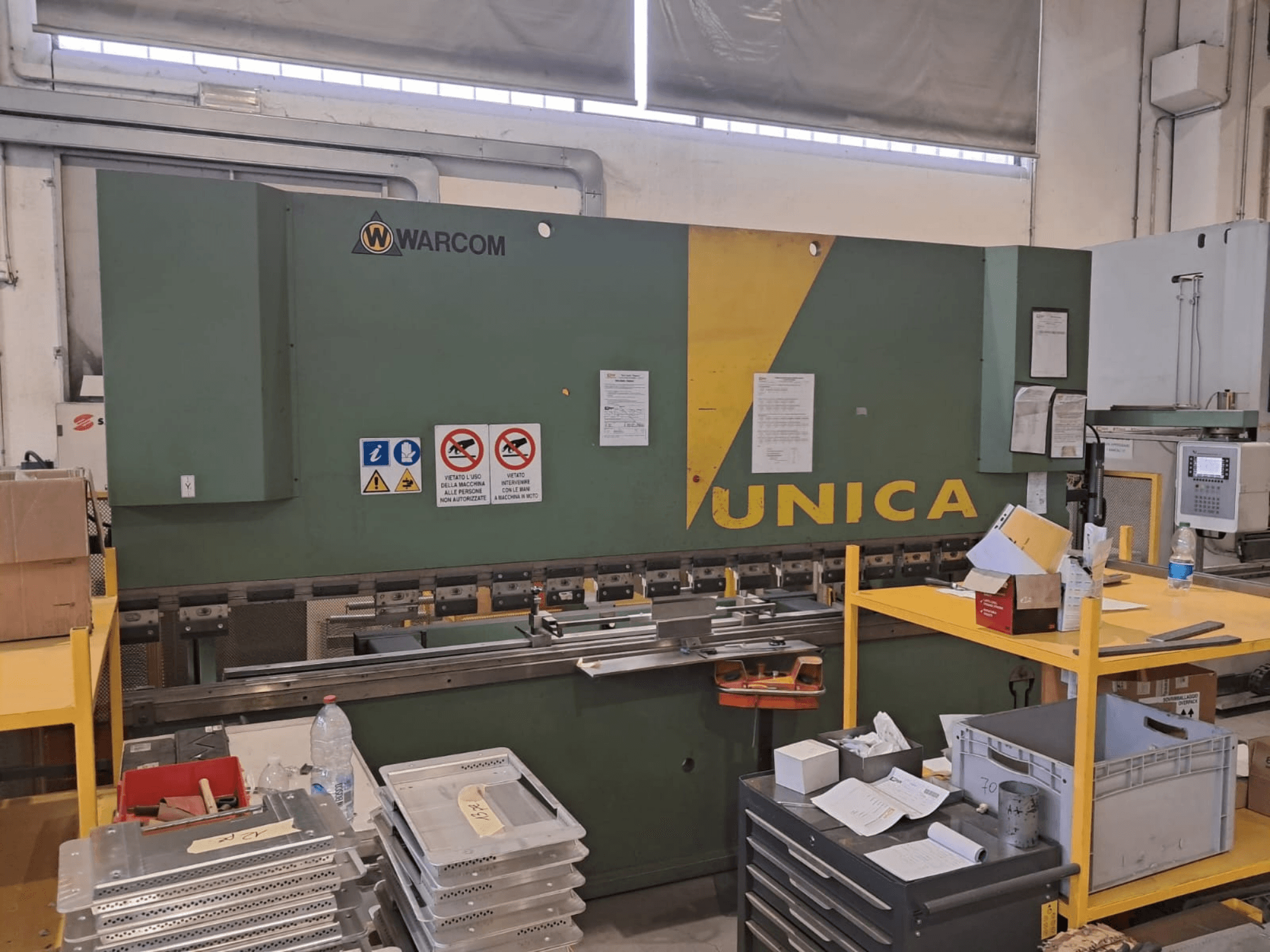 Hydraulický lis Warcom UNICA 100-40, pohled zepředu, zelené tělo se žlutými prvky, viditelné bezpečnostní značky, nástroje a materiály na stole.