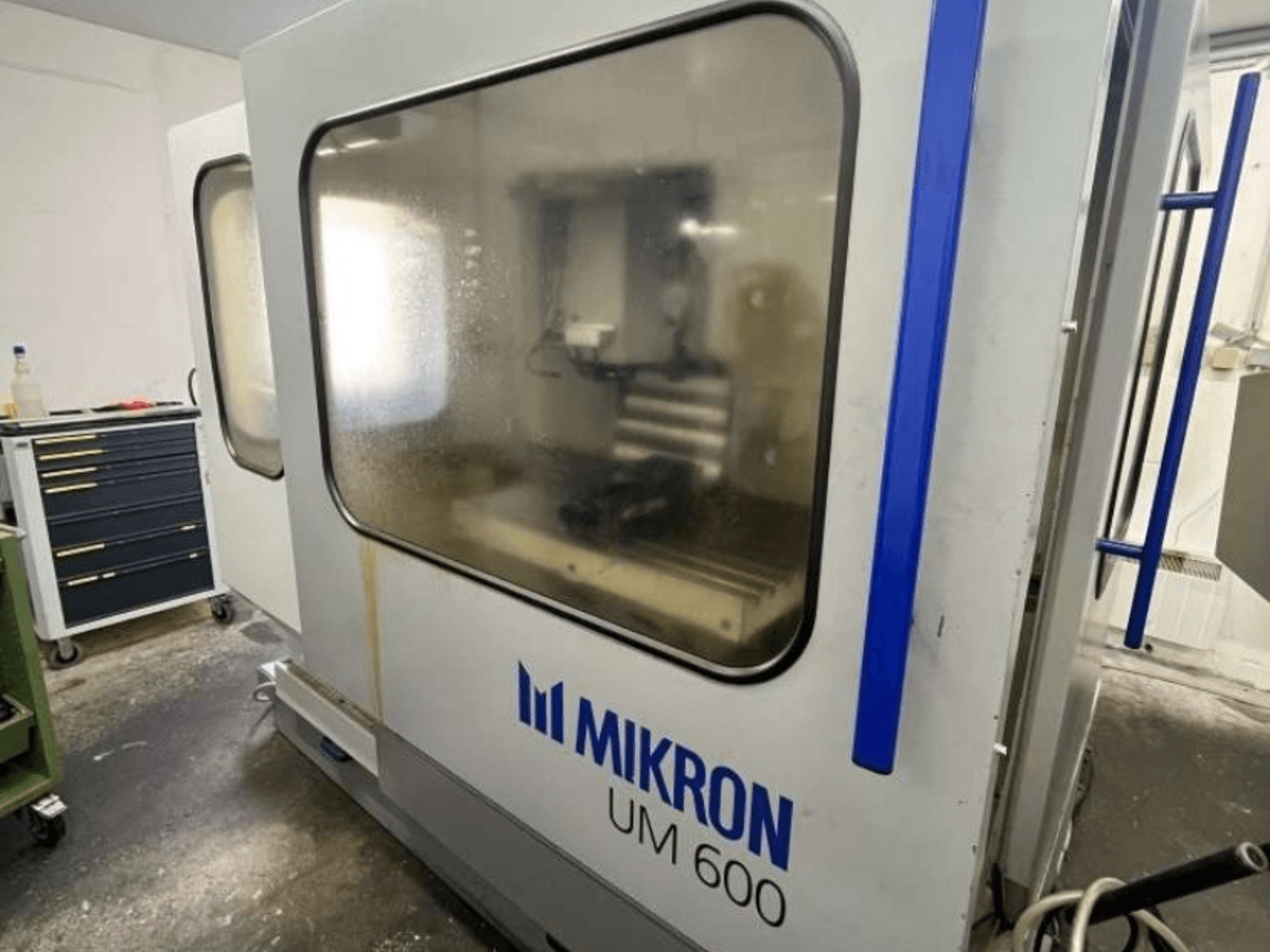 Univerzální obráběcí centrum MIKRON UM 600 TNC 410/426, boční pohled, se zamlženým sklem a viditelným úložným prostorem pro nástroje v blízkosti.