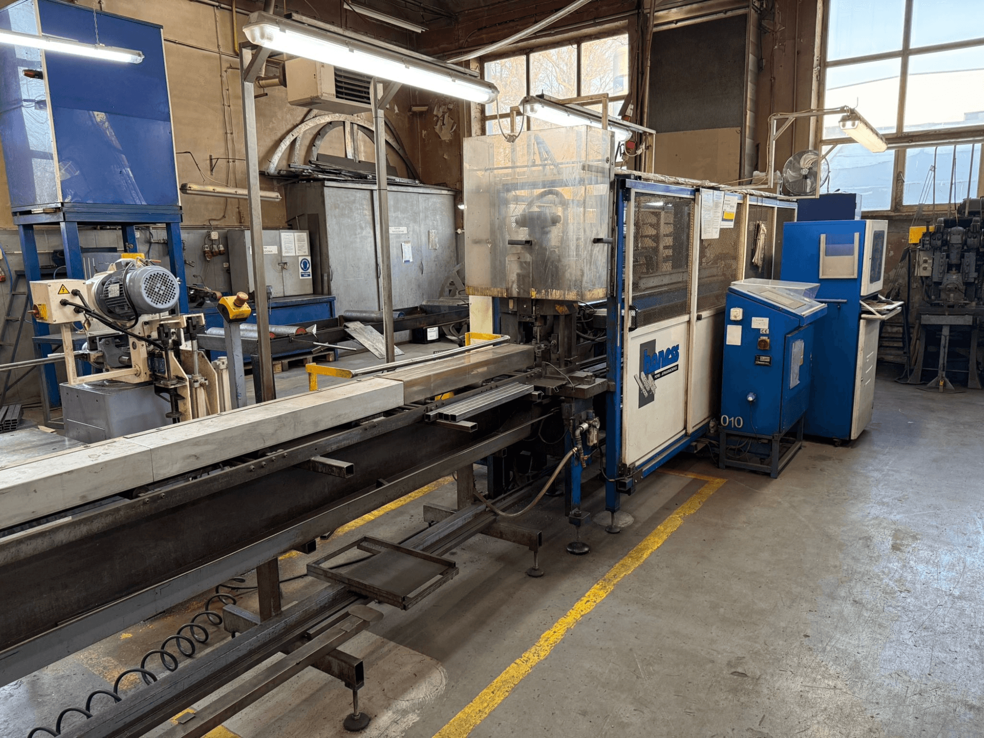 Průmyslová dílna s modrým strojem Haweas, boční pohled na dopravník a zařízení ve velkém, dobře osvětleném prostoru.