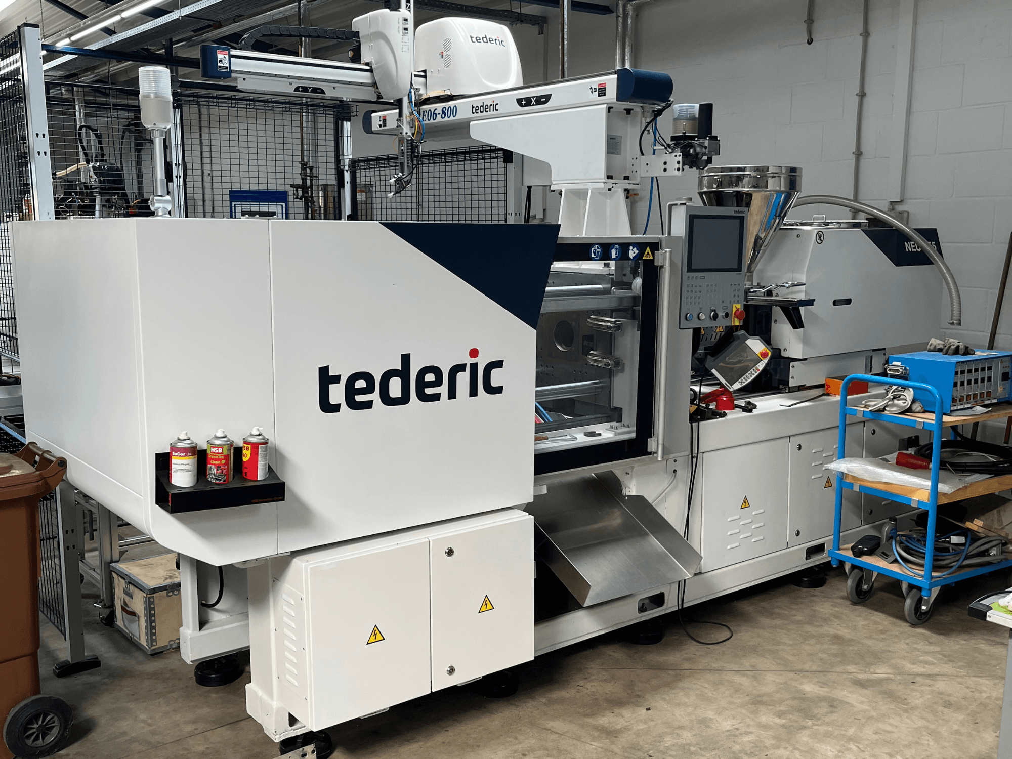 Hydraulický vstřikovací stroj TEDERIC NEO.E55/e110h v dílně, boční pohled s ovládacím panelem a přilehlými nástroji.