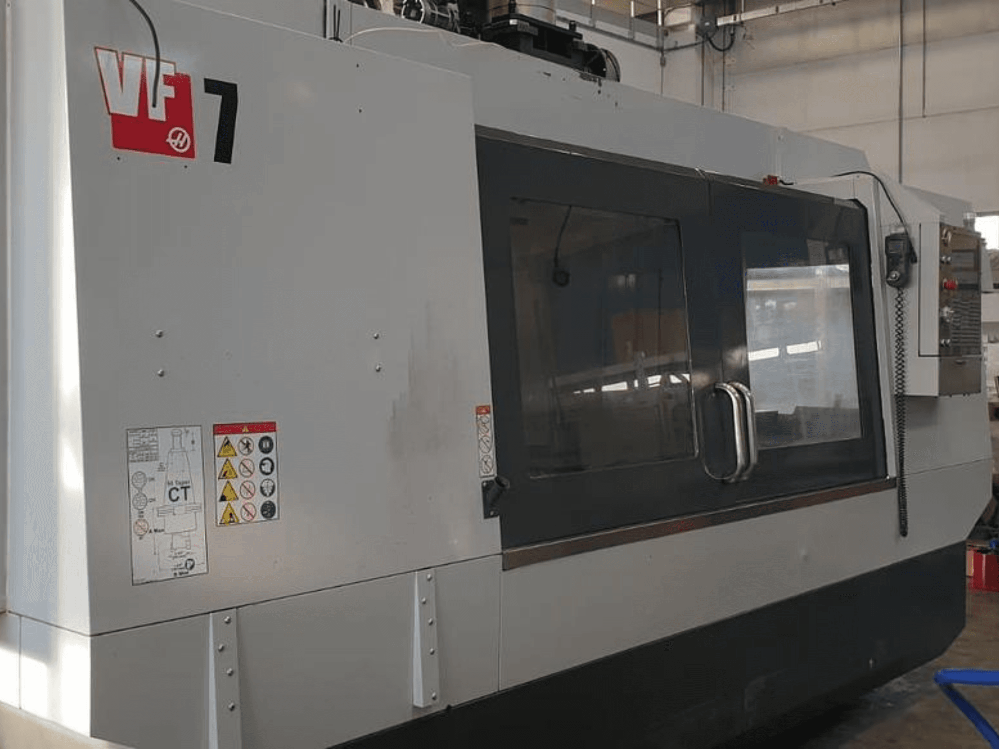 CNC stroj Haas VF-7 v bočním pohledu, s kovově šedým povrchem, ovládacím panelem a bezpečnostními štítky.