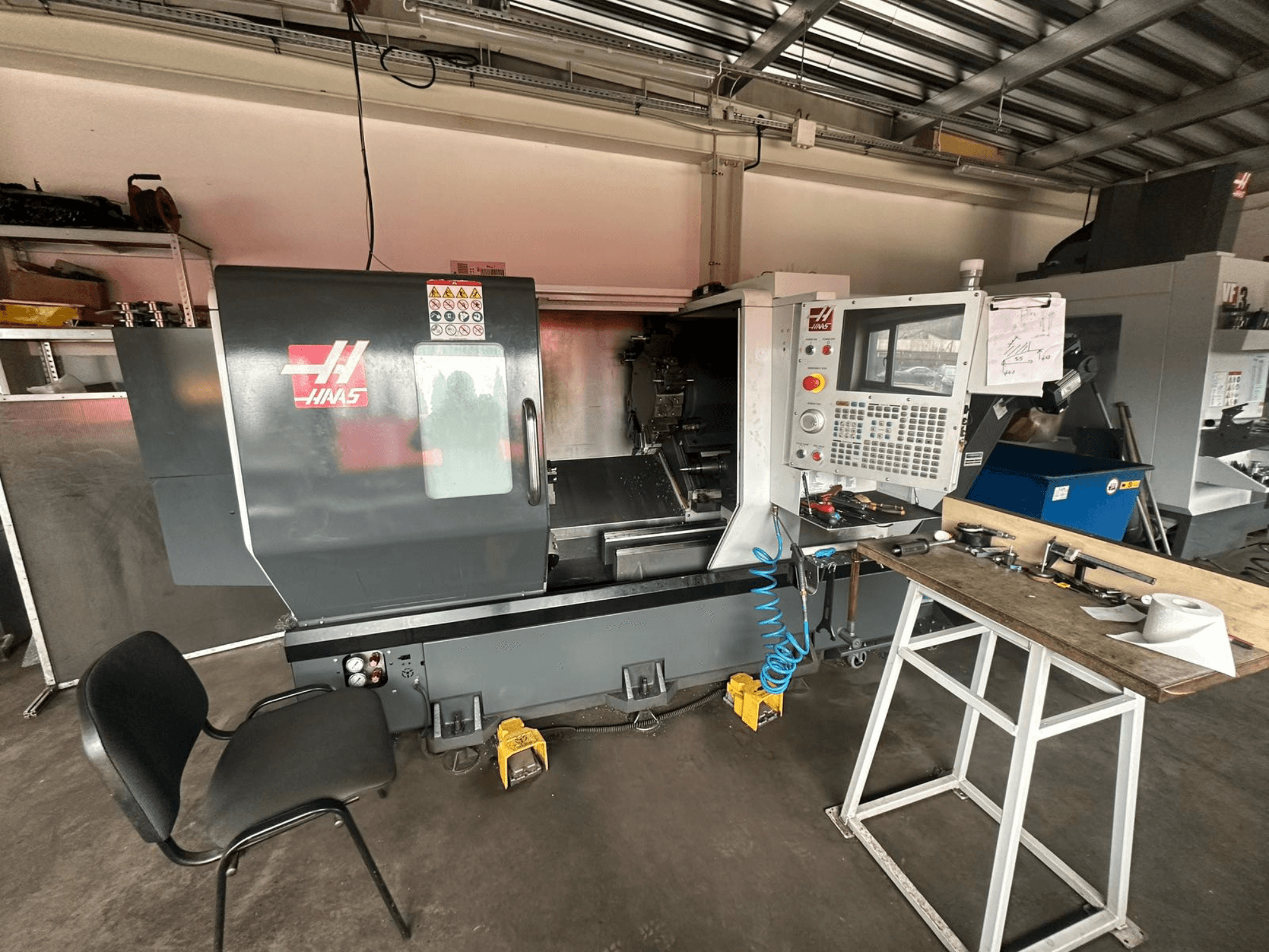 CNC soustruh Haas (model ST-10) v dílně, boční pohled ukazuje ovládací prvky, nástroje na pracovním stole a židli poblíž.
