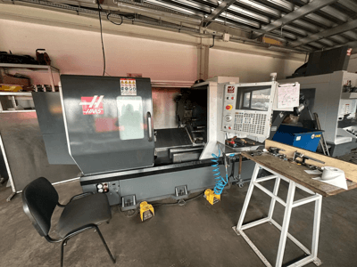 CNC soustruh Haas (model ST-10) v dílně, boční pohled ukazuje ovládací prvky, nástroje na pracovním stole a židli poblíž.