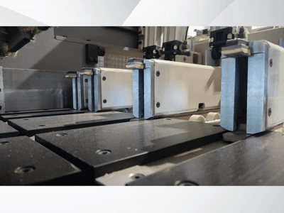 Pohled zblízka na upínací mechanismus přesného stroje s kovovými čelistmi, s černými pracovními plochami a viditelnými vodicími lištami.