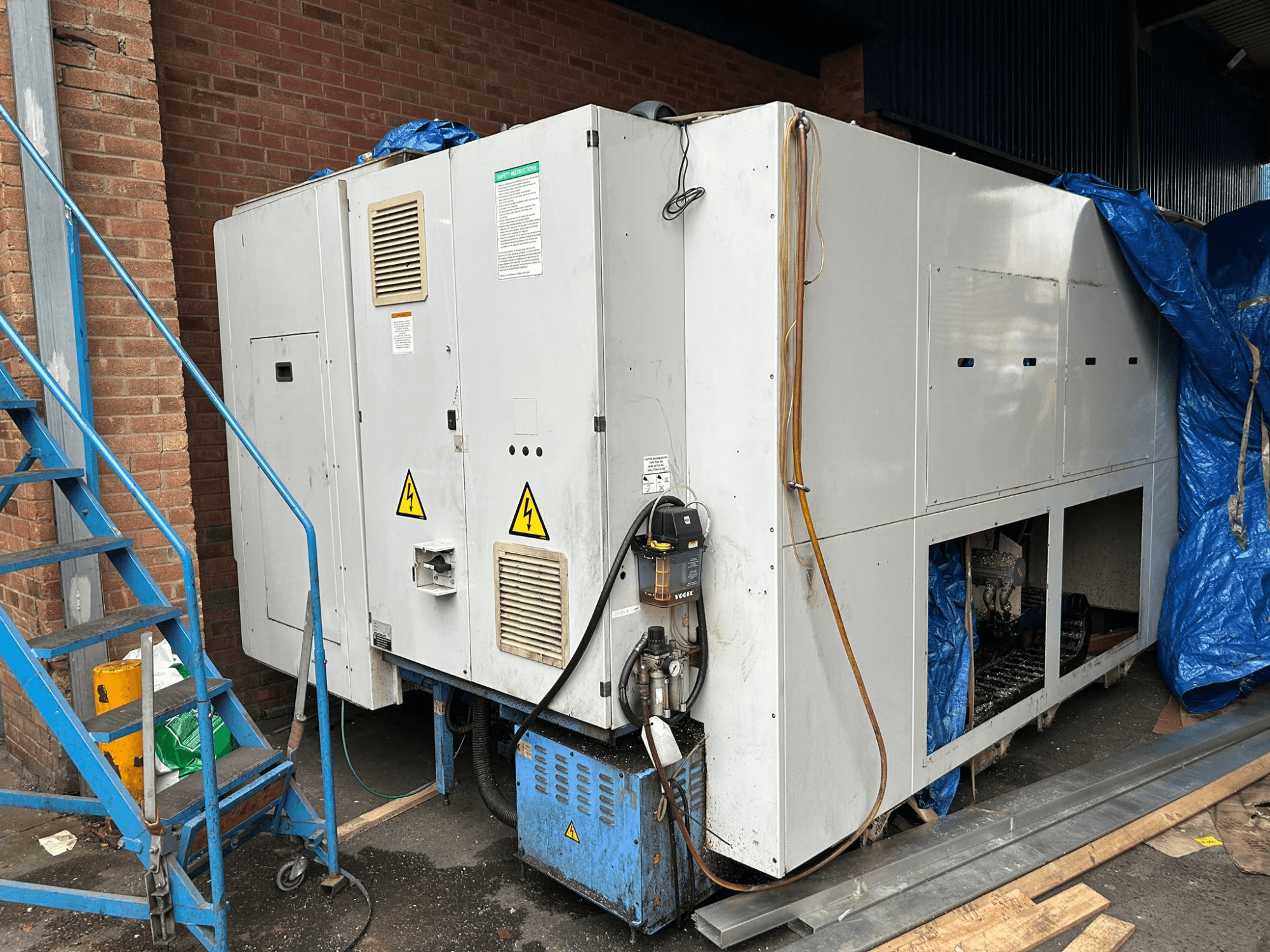 Boční pohled na velkou průmyslovou chladicí jednotku, značka a model nejasné, s elektrickými přípojkami, výstražnými značkami a modrým plachtovým krytem.