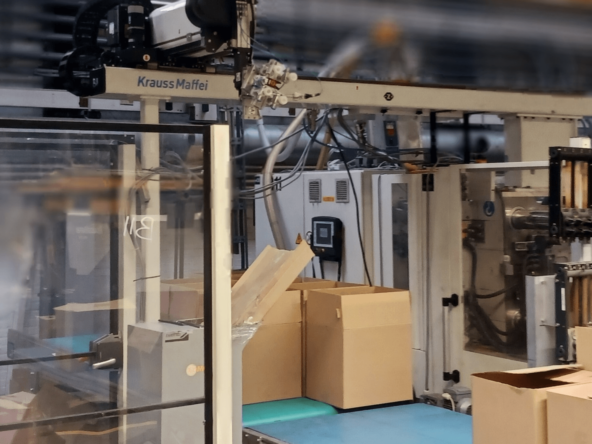 Automatizační systém Krauss Maffei v továrně, pohled zepředu ukazuje krabice na dopravníku a strojní součásti v provozu.
