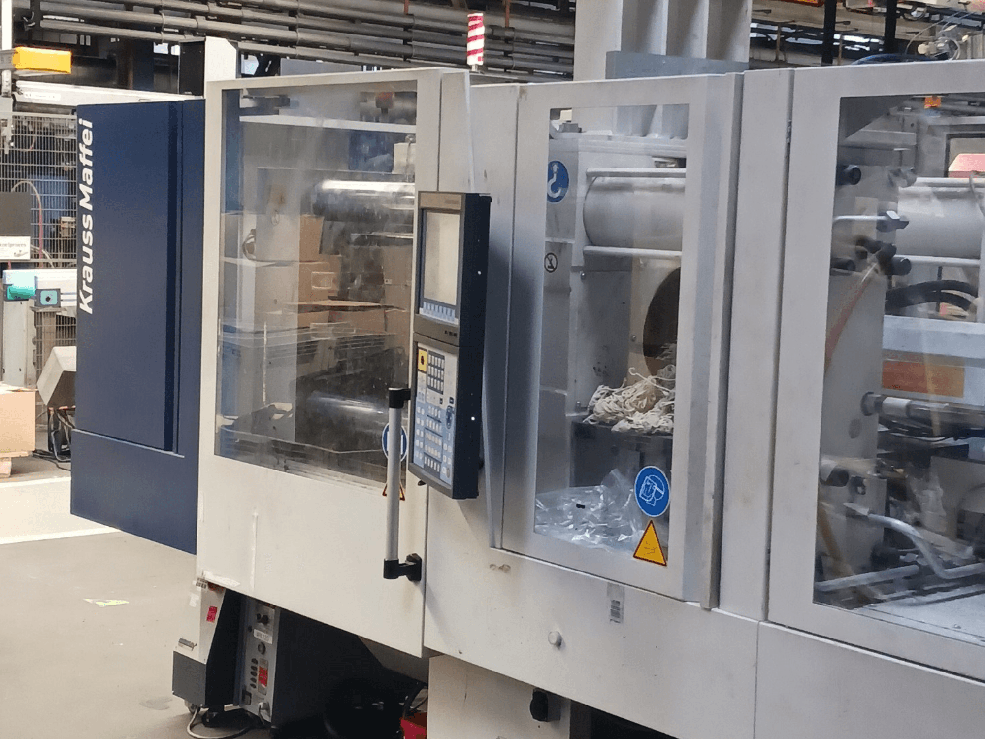 Vstřikovací stroj KraussMaffei, boční pohled, zobrazující ovládací panel, strojní zařízení a obalové materiály v pozadí.