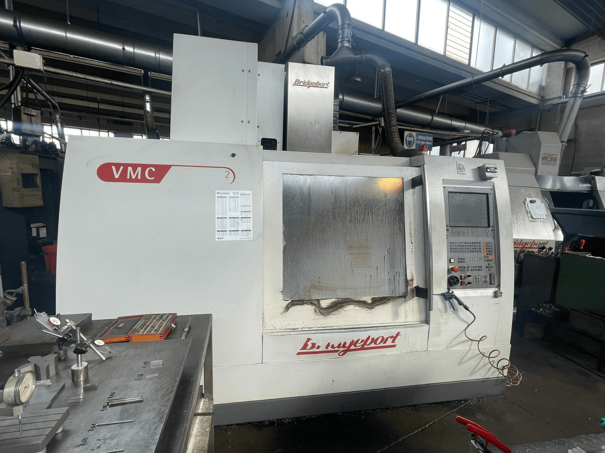 Vertikální obráběcí centrum Bridgeport VCM 800-30 v dílně, s ovládacím panelem a stolem na nástroje. Pohled zepředu s viditelnými detaily.